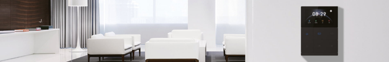 Modernes, hell eingerichtetes Wohnzimmer mit weißen Sofas, großen Fensterfronten und dezenten Vorhängen. An der rechten Wand ist das Touch+ Pad der GVS Walz Serie montiert. Das schwarze Touchpanel mit Display zeigt Uhrzeit, Wetterinformationen und verschiedene Smart-Home-Funktionen zur Steuerung von Beleuchtung, Klima und weiteren Gebäudefunktionen. Die Aufnahme verdeutlicht die elegante Integration des Bedienpanels in ein hochwertiges Wohnambiente.